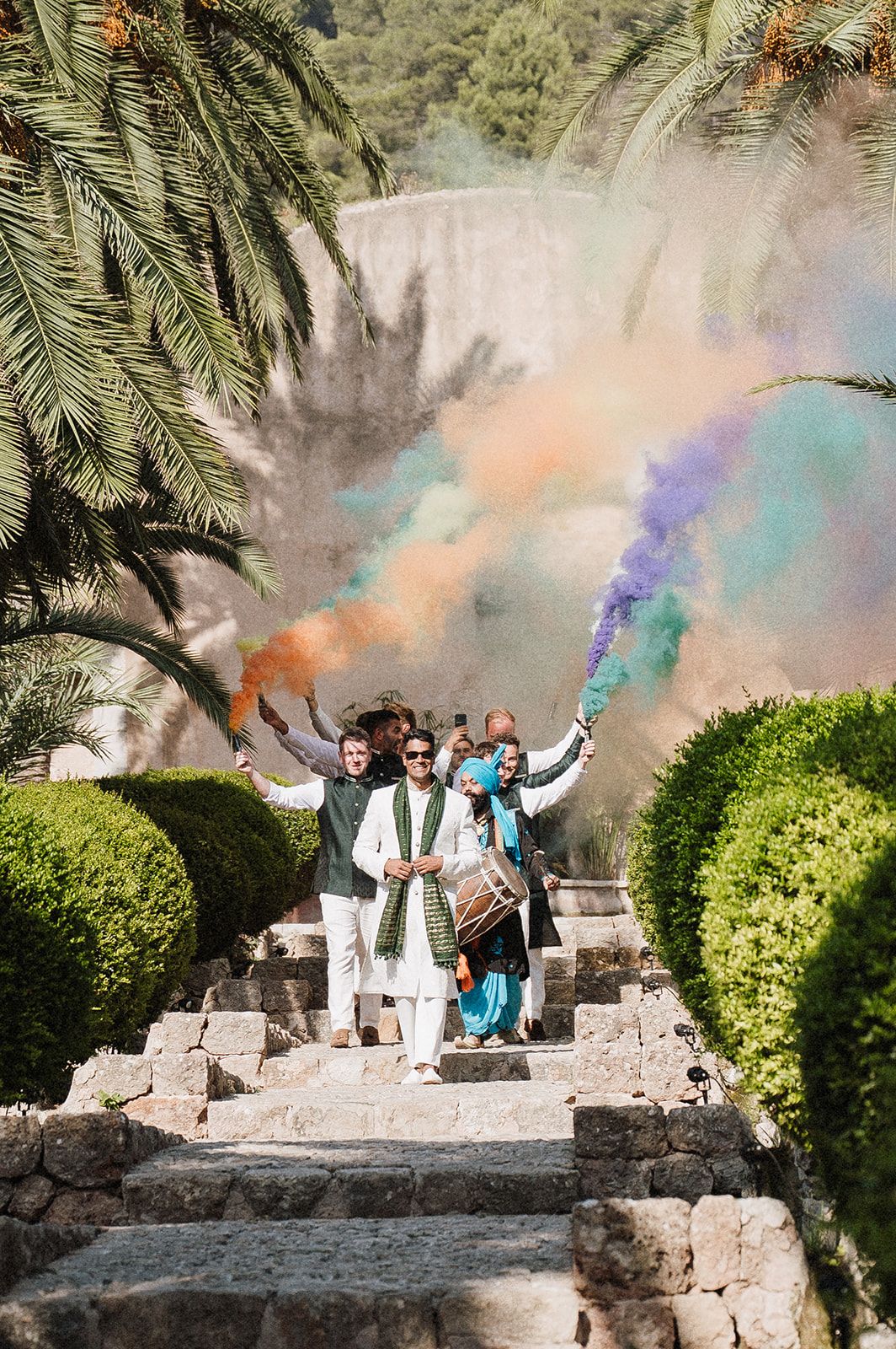 Groomsmen celebrating with colorful smoke bombs on stone steps at Indian wedding.