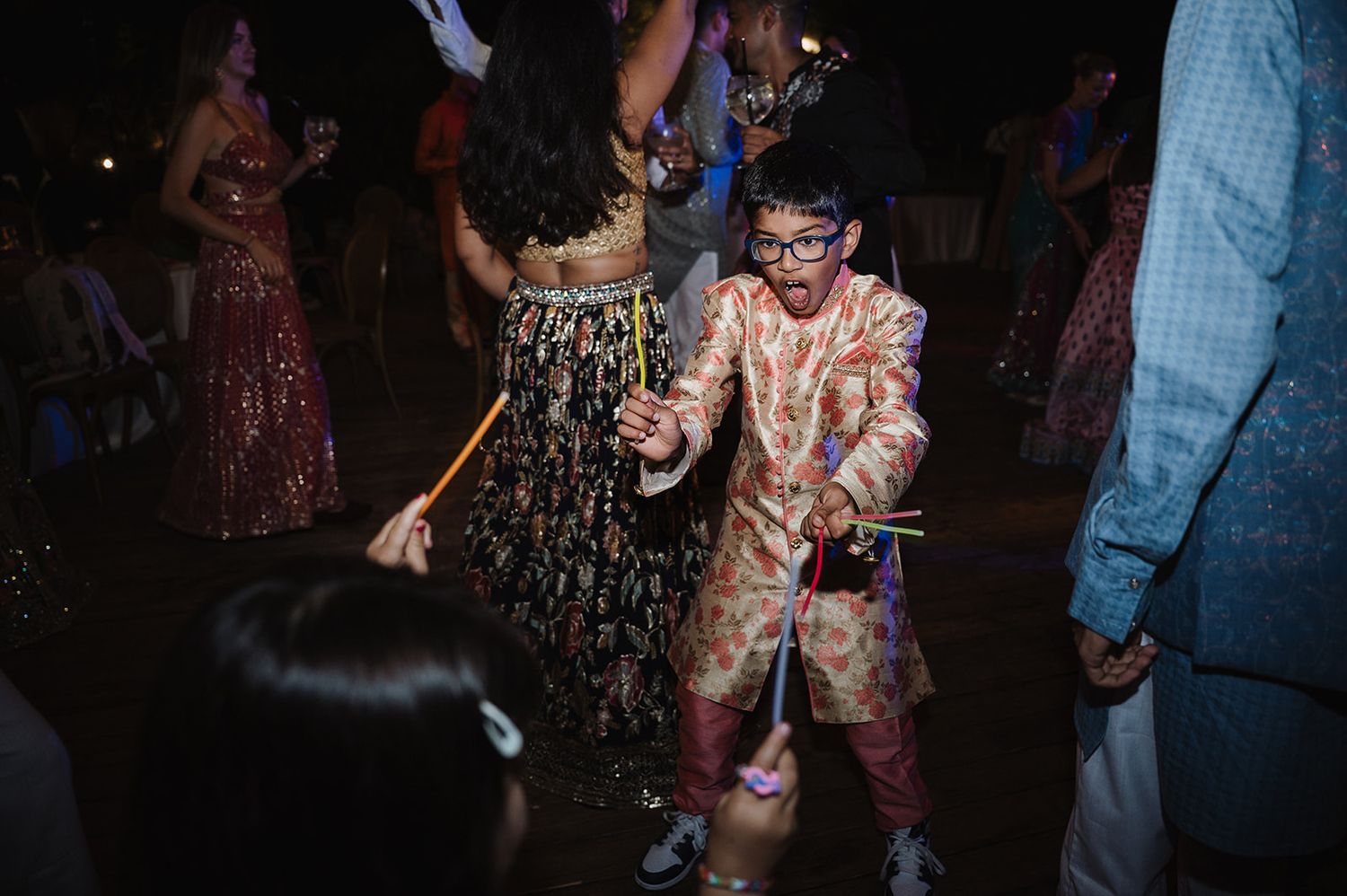 Young boy in pink embroidered sherwani dances joyfully with glow sticks at an Indian wedding reception.