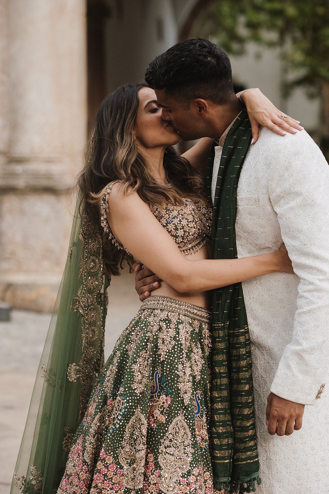 Couple sharing a romantic kiss in traditional Indian wedding attire, green dupatta and embroidered lehenga.