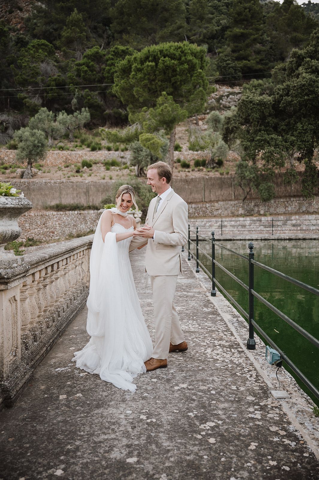 Bride and groom share private moment on stone balustrade walkway beside green water at historic estate.