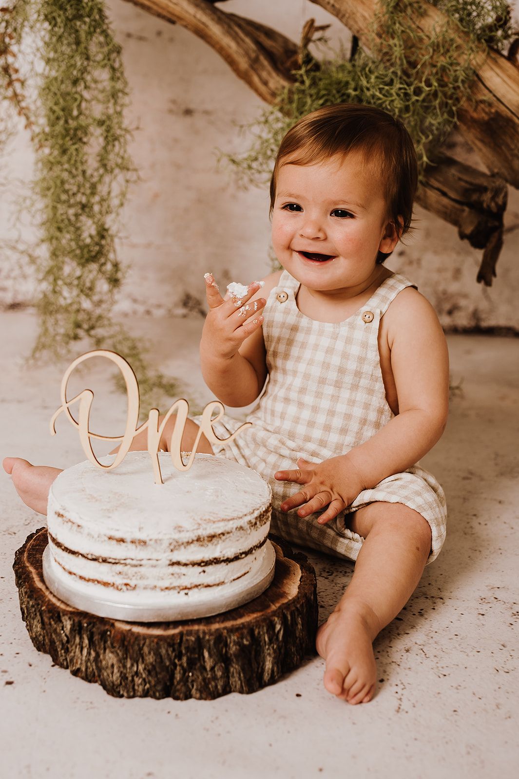 Smiling baby in gingham romper celebrating first birthday with a naked cake topped with 
