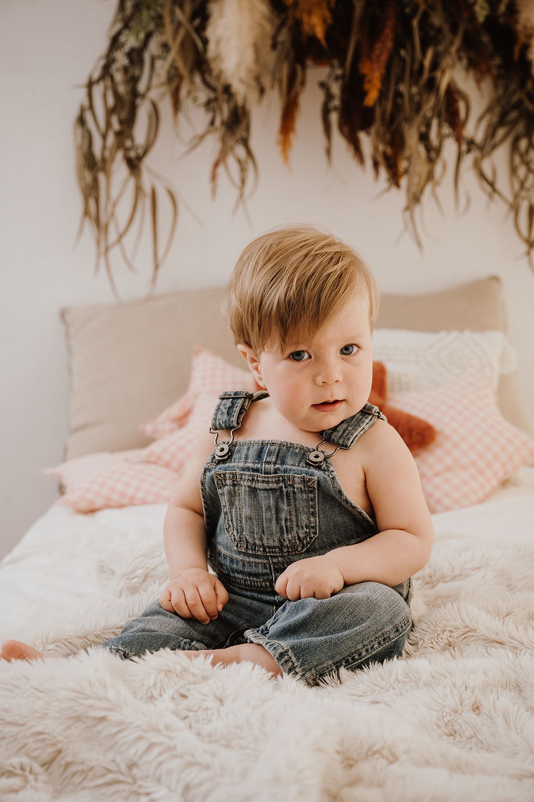 Cute toddler in denim overalls sitting on a fluffy white bed with dried botanicals overhead.
