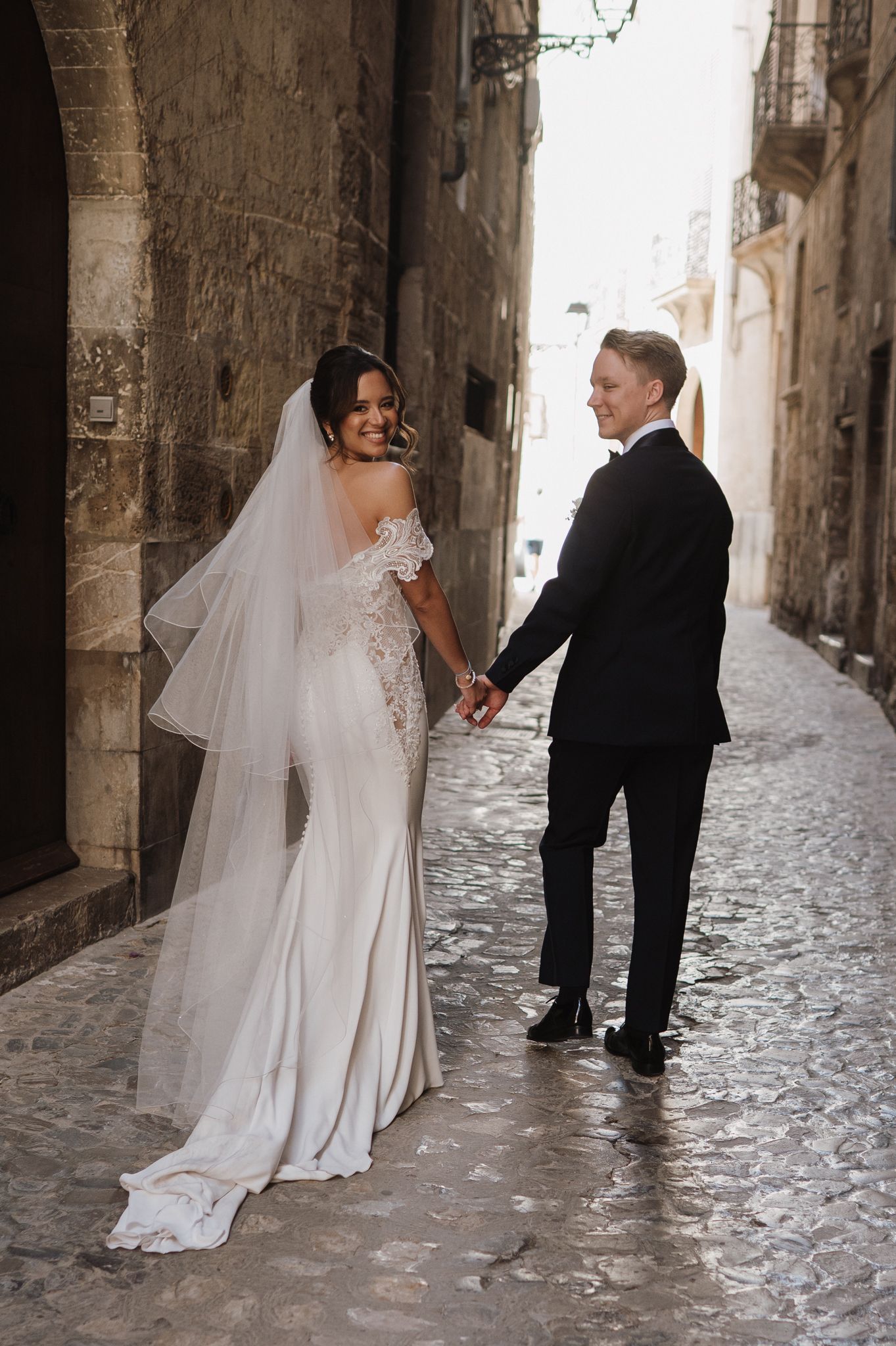 Bride and groom holding hands walking cobblestone alley in Sicily in elegant wedding attire.