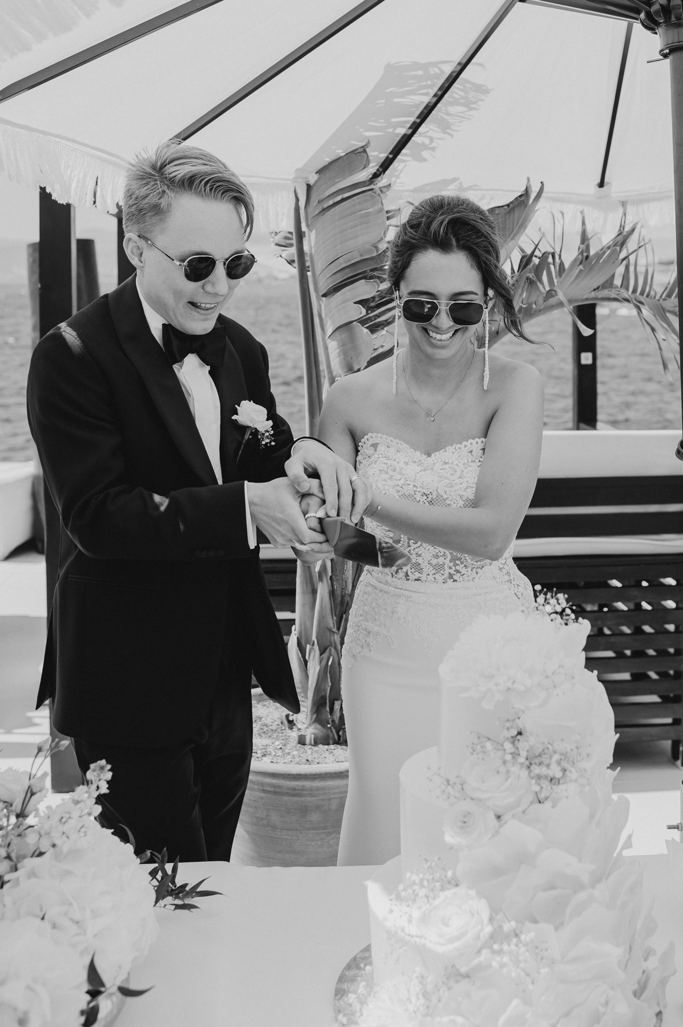 Bride and groom wearing sunglasses cut wedding cake together in fun black and white photo.