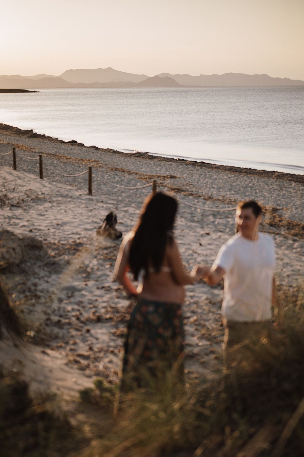 Couple holding hands on a sandy beach path at sunset with mountains and ocean visible in the background.