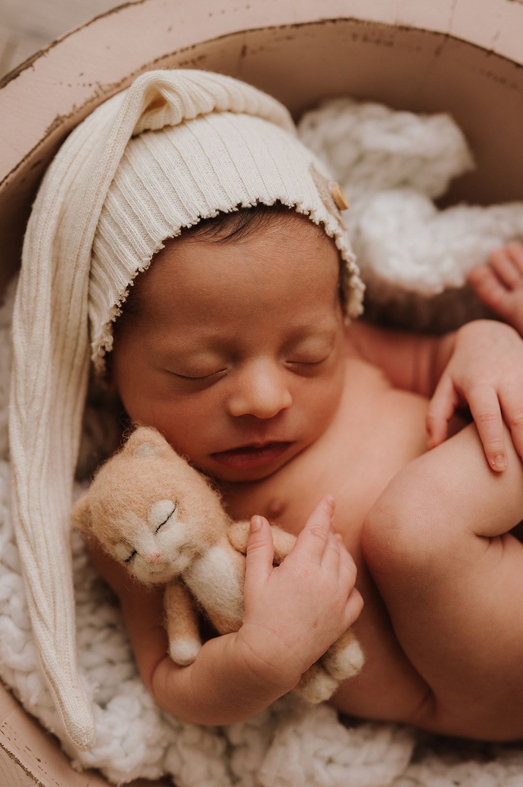 Sleeping newborn baby in cream knit hat cuddles a tiny teddy bear in a round wooden bowl.