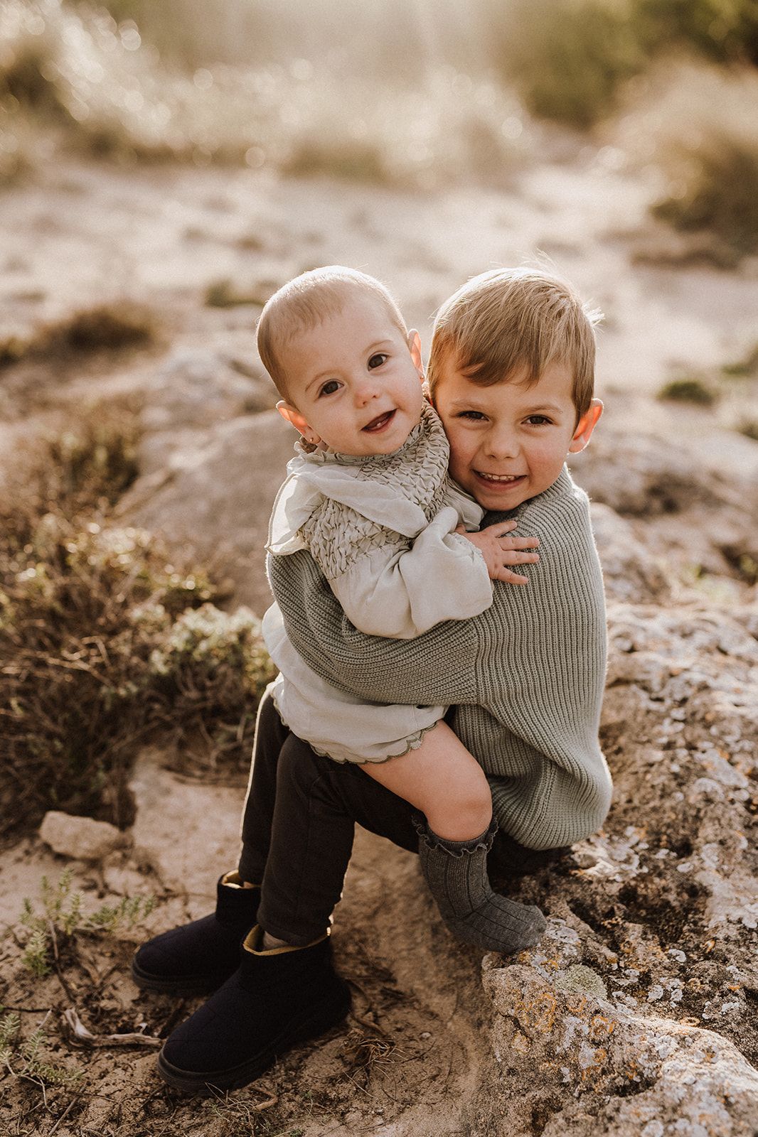 Two young siblings hugging outdoors on rocky terrain in warm golden light, wearing cozy knit sweaters.