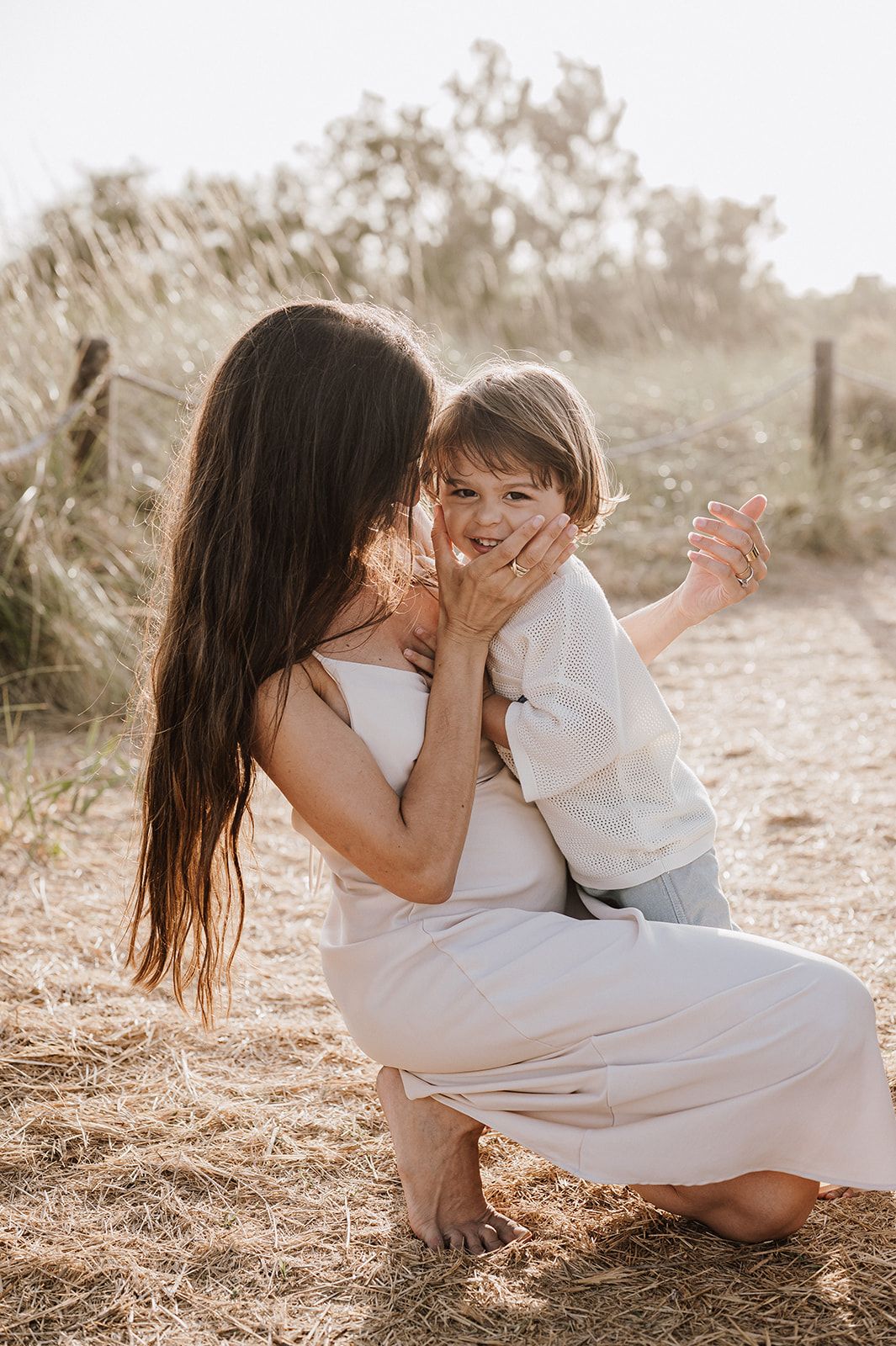 A barefoot pregnant woman in white dress gently cups the face of her smiling toddler outdoors.