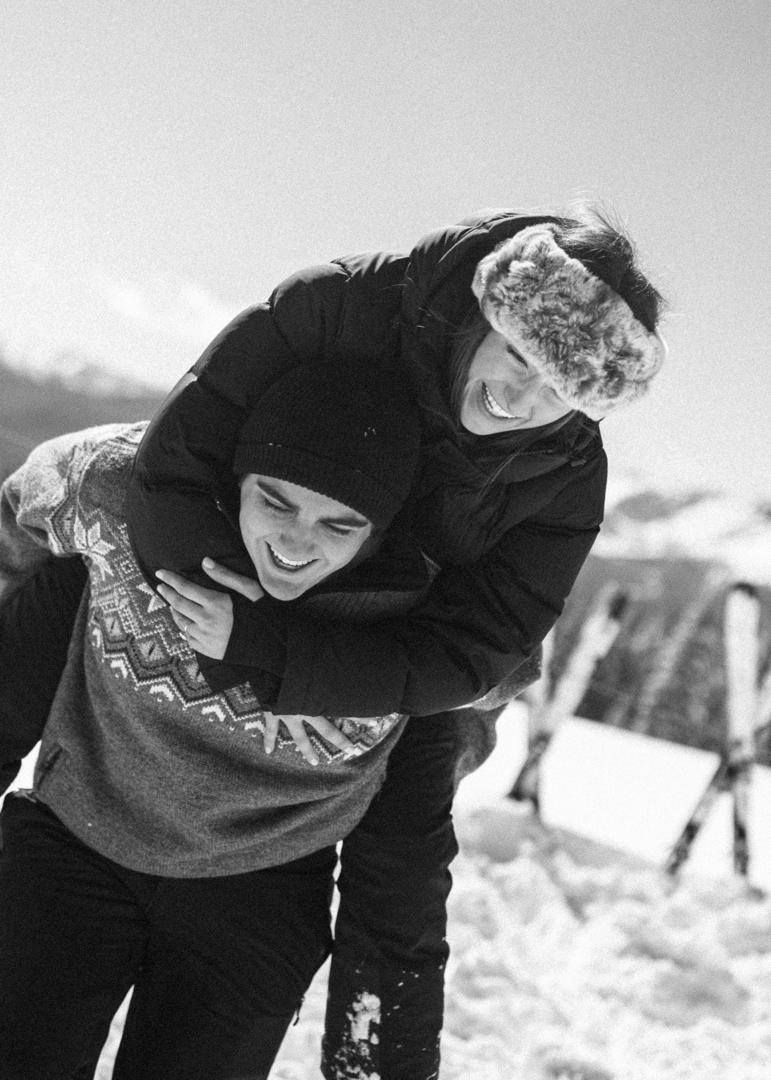 Playful couple laughing in black and white photo with woman hugging man from behind in snow.