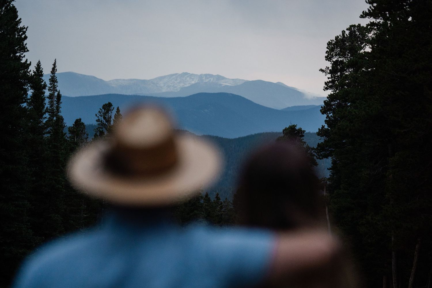 A couple stands together gazing at a stunning panoramic mountain vista through tall pine trees at dusk.
