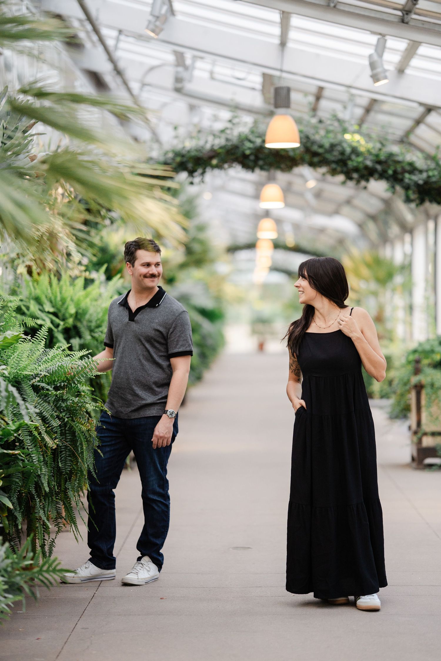 Couple smiling and walking together through a lush greenhouse corridor filled with tropical ferns and plants.