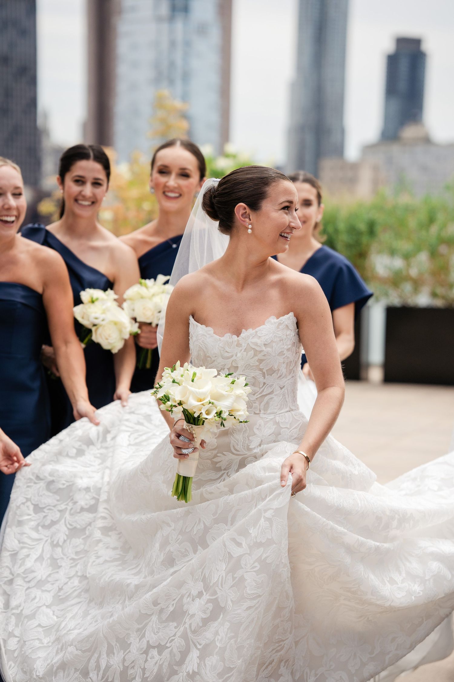 Smiling bride in strapless lace gown twirls with bouquet surrounded by navy-dressed bridesmaids on rooftop.