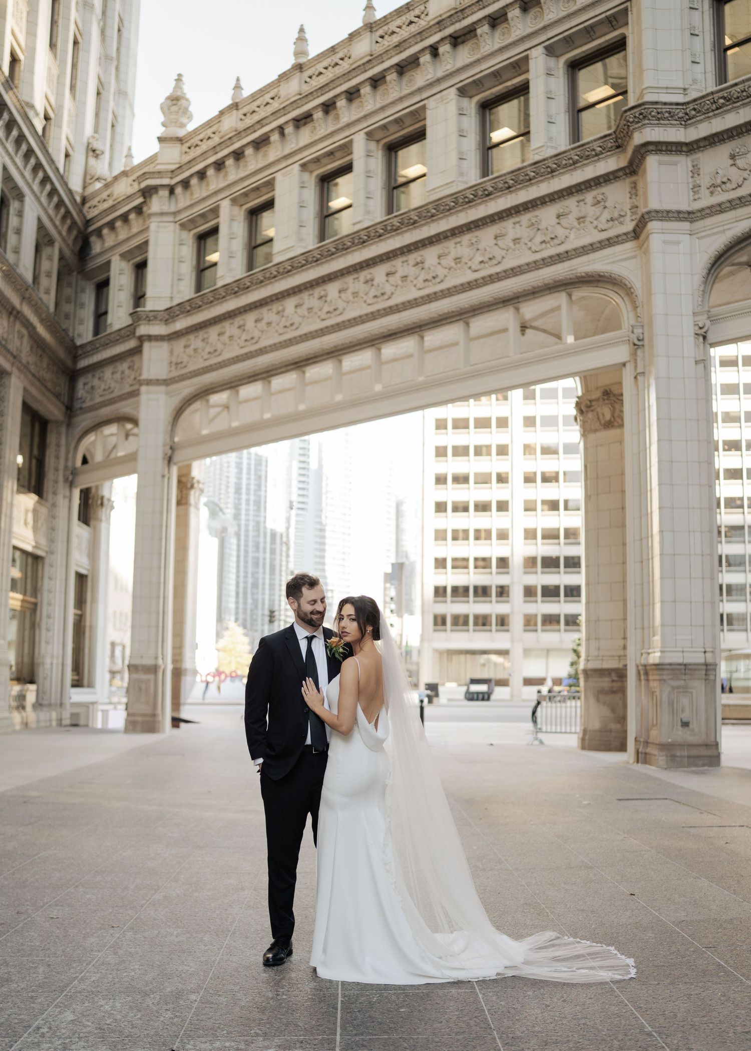 Bride and groom hold hands under ornate stone arch in Chicago's Wrigley Building plaza on their wedding day.