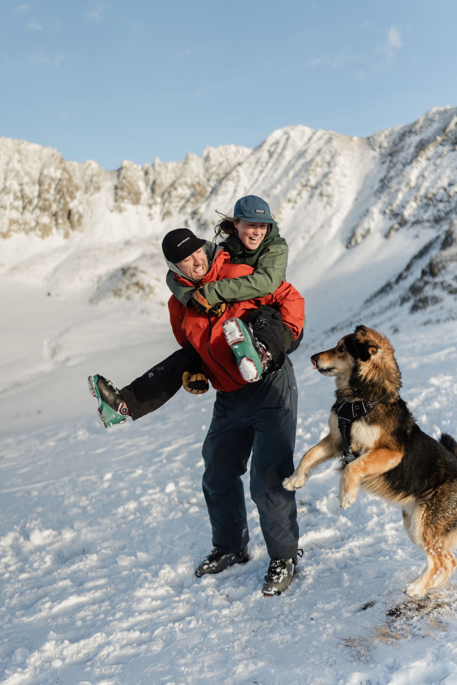 A man in a red jacket lifts a laughing woman while their large dog jumps playfully in a snowy mountain setting.