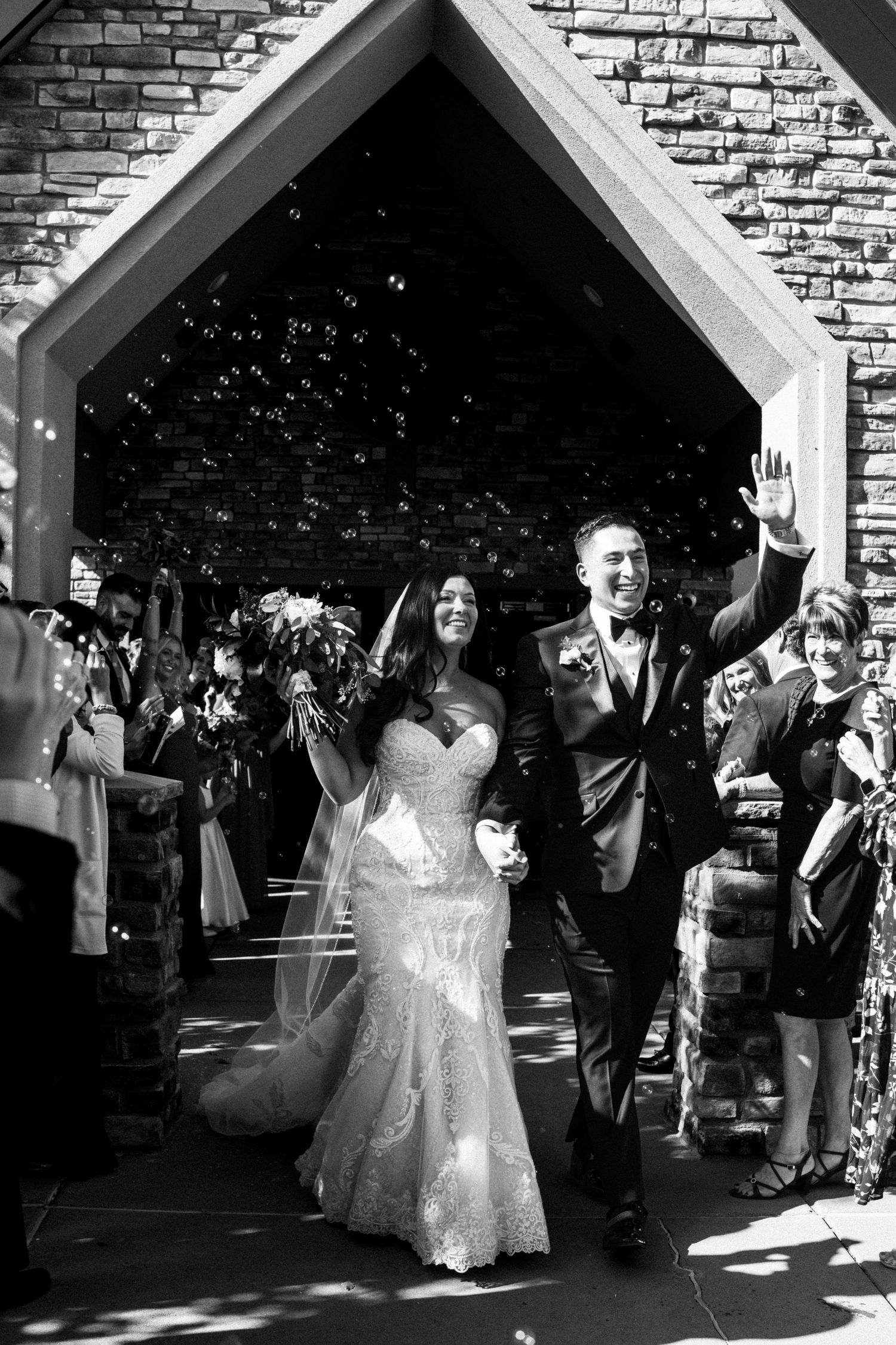 Newlyweds celebrating joyful exit from stone church surrounded by bubbles and guests in black and white.