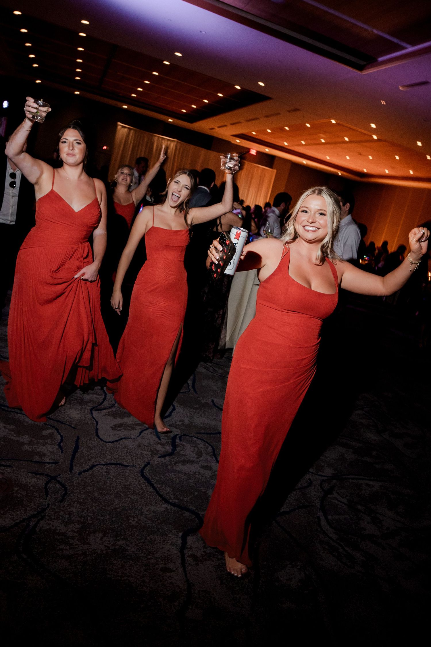 Three bridesmaids in red gowns dancing and celebrating on wedding reception dance floor.