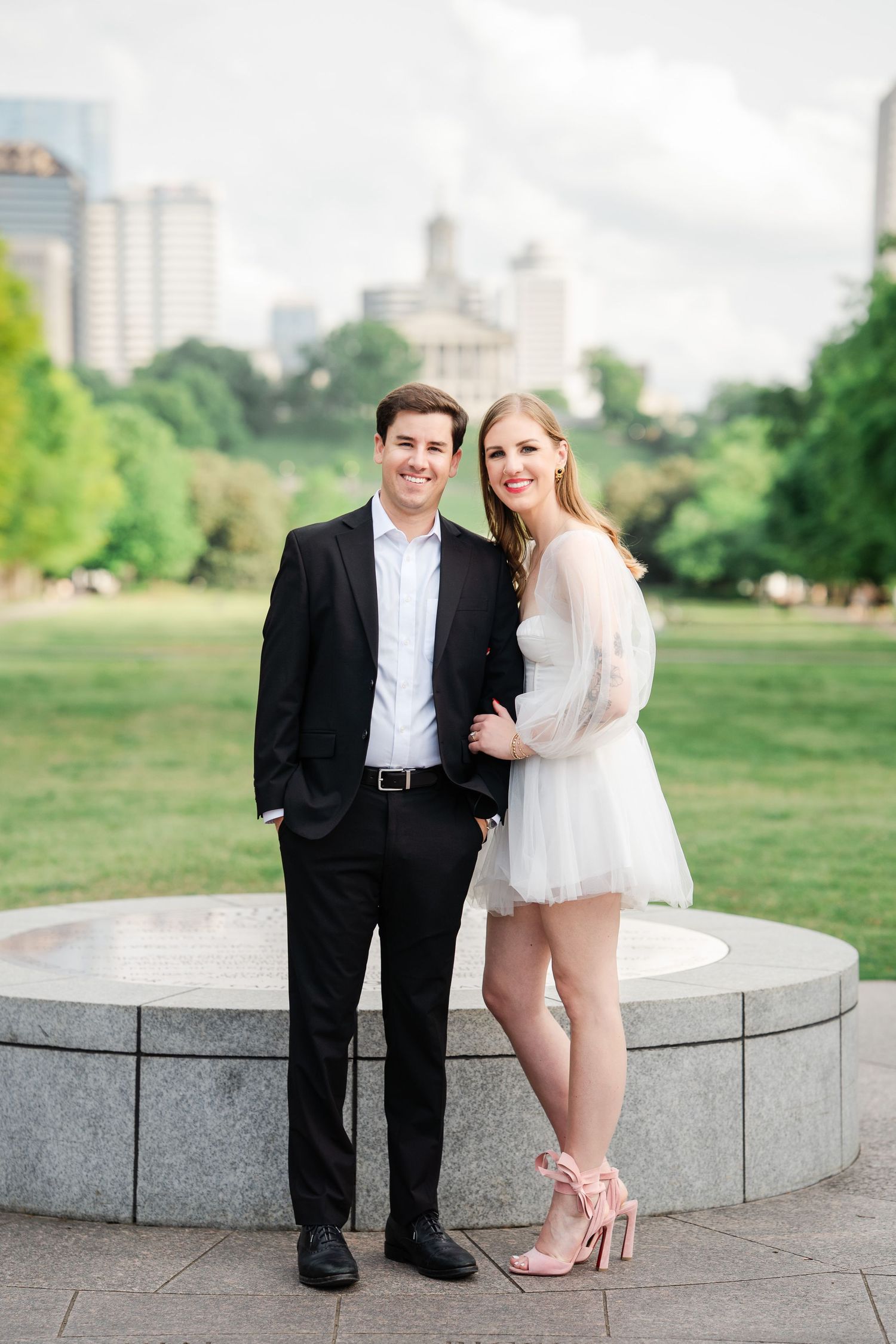 Smiling couple posing at outdoor fountain with city skyline, man in black suit and woman in white mini dress.