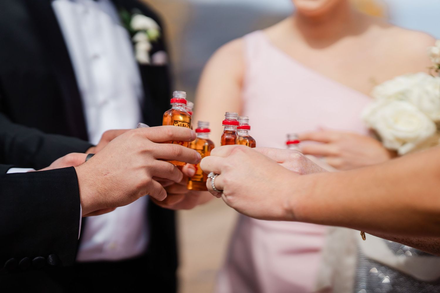Wedding party clinking mini liquor bottles together in colorful celebratory toast during outdoor ceremony.