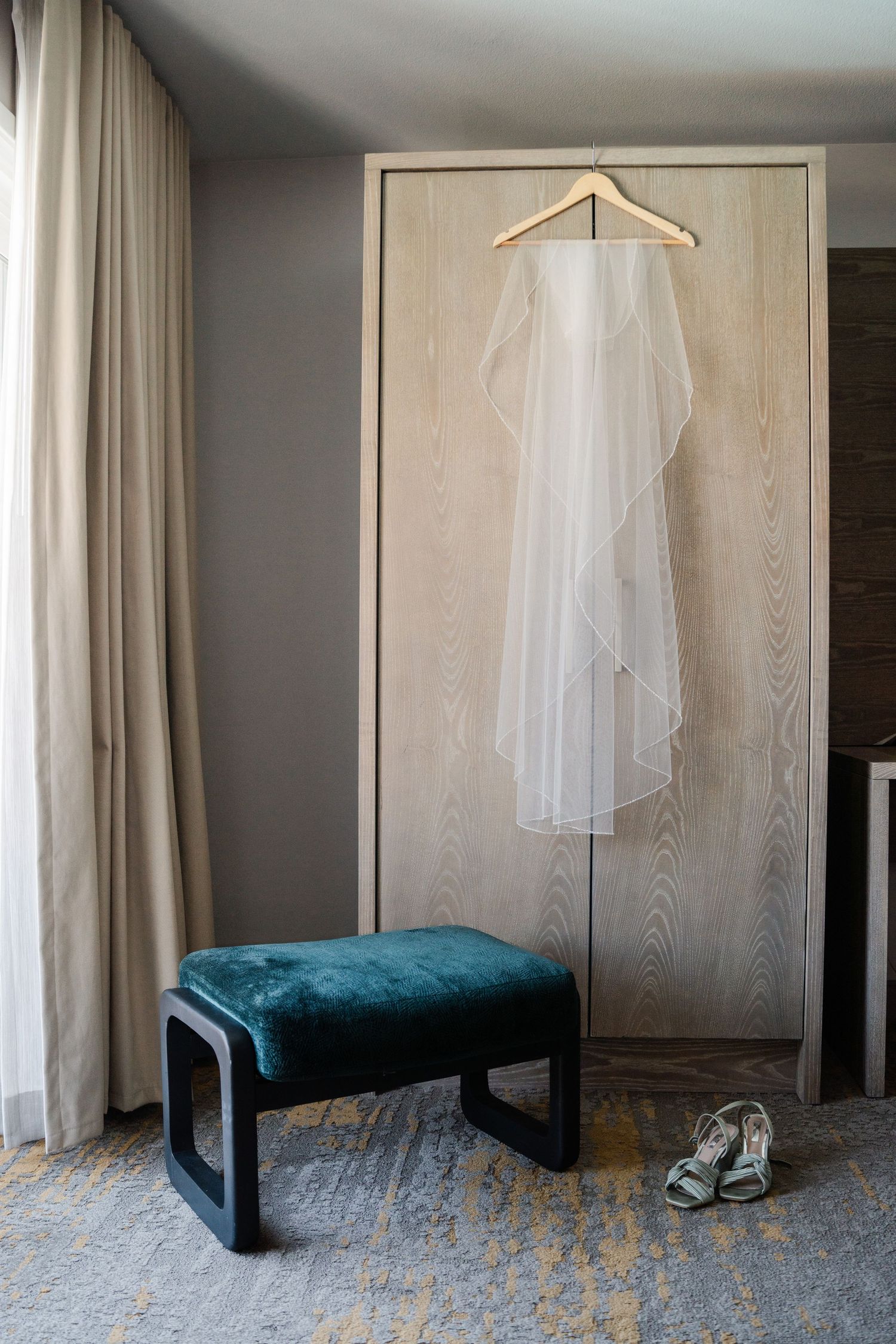 White wedding veil hanging on a wooden wardrobe with silver heels on the floor beside a velvet stool.