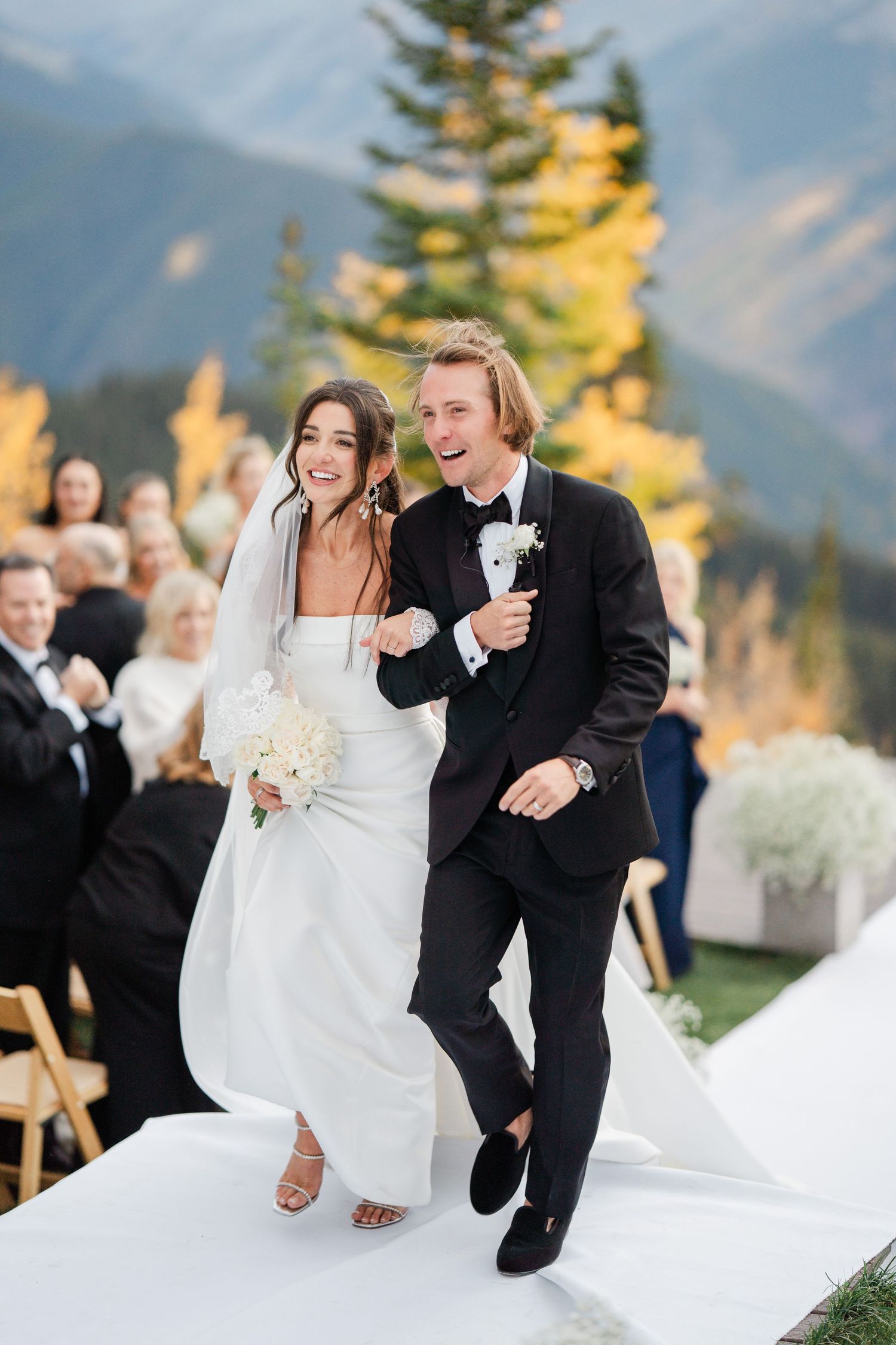 Ecstatic newlyweds celebrating as they walk down white aisle with autumn mountain backdrop.