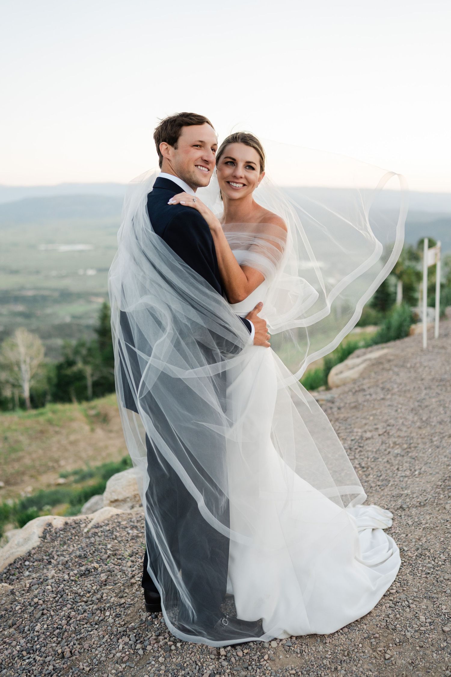 Bride and groom embracing with veil flowing in the wind at a scenic mountain overlook.