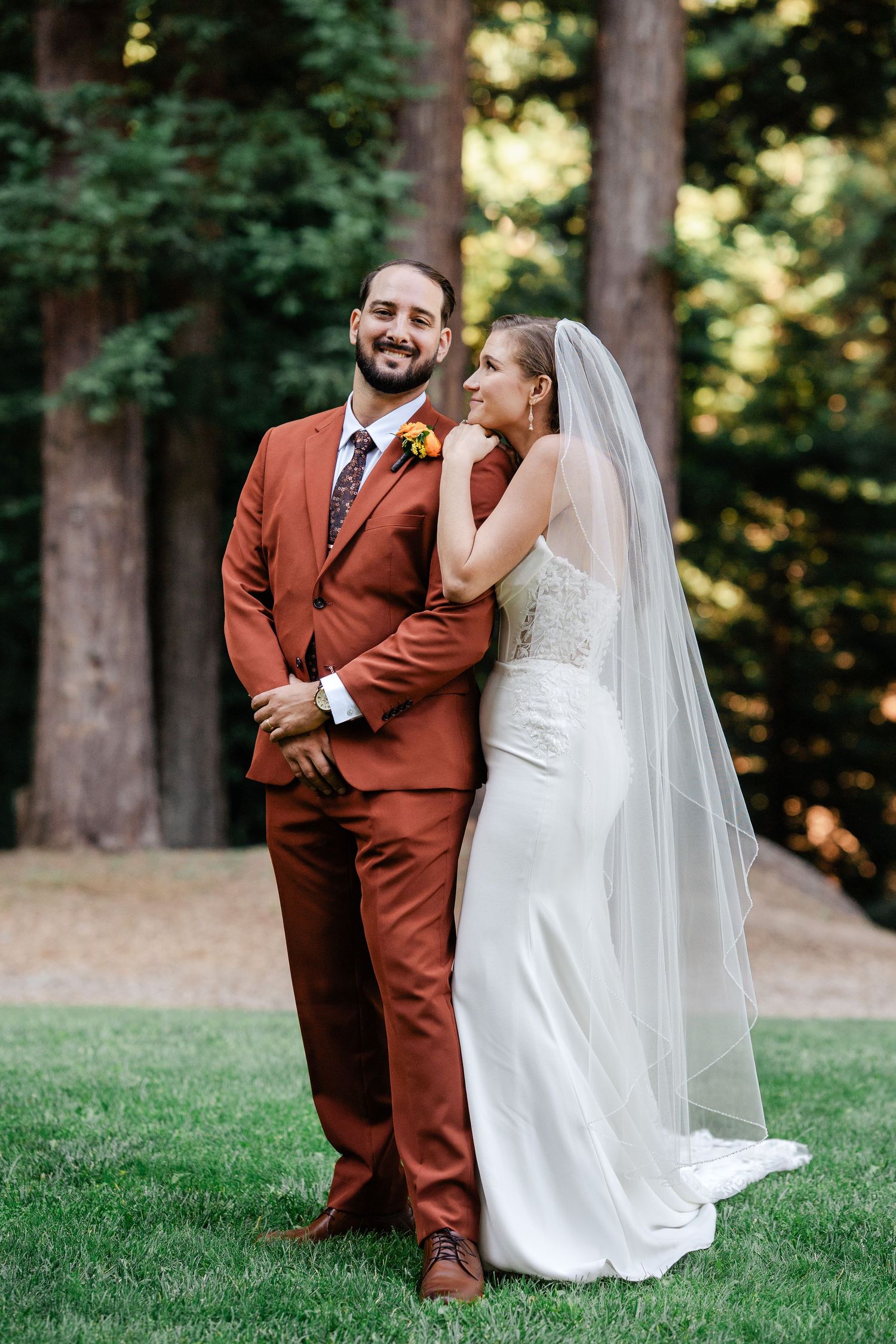 Bride in white lace gown with veil and groom in rust suit posing in redwood forest.