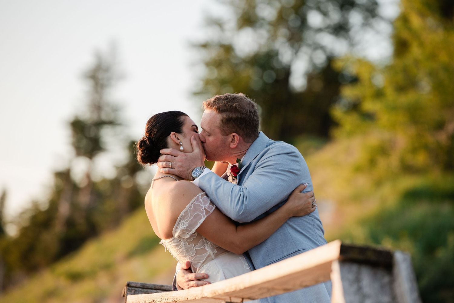 A bride and groom share a romantic kiss outdoors at golden hour with lush green trees in the background.