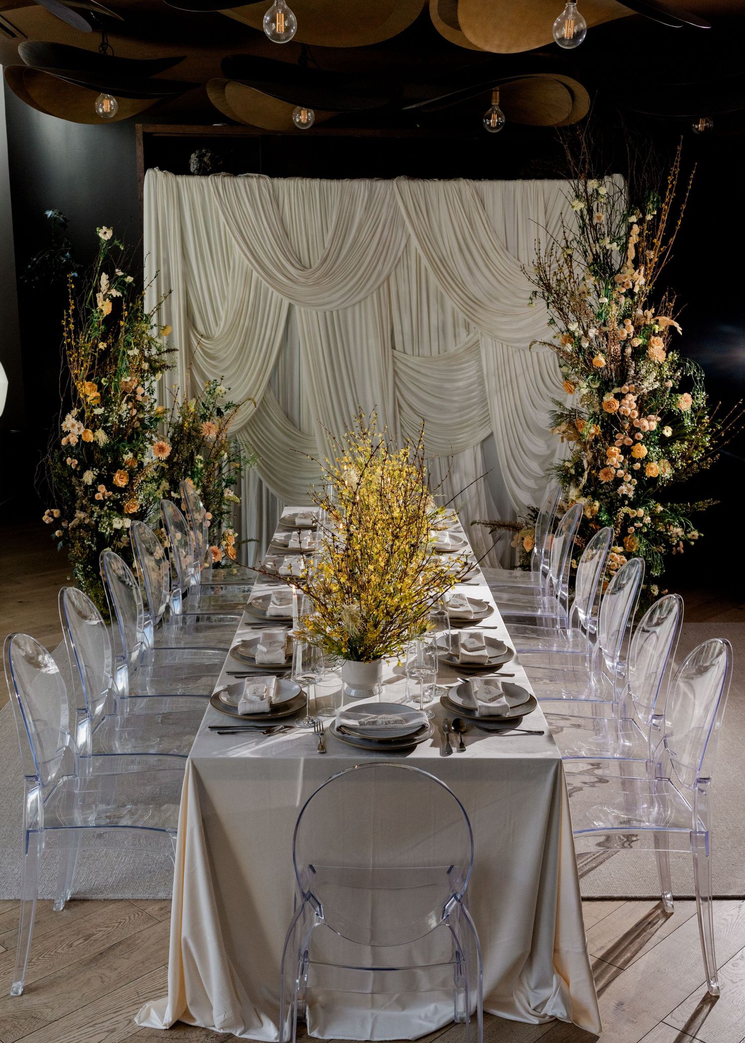 Elegant wedding reception table with ghost chairs, floral arrangements, and a draped white backdrop in a modern venue.