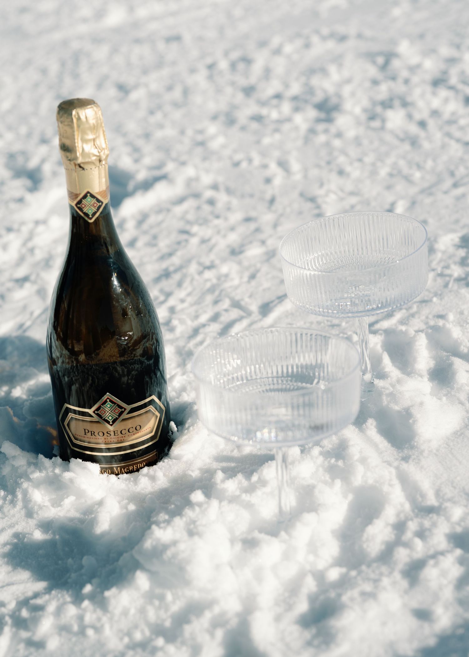 Bottle of Prosecco chilling in fresh snow beside a crystal coupe glass outdoors.