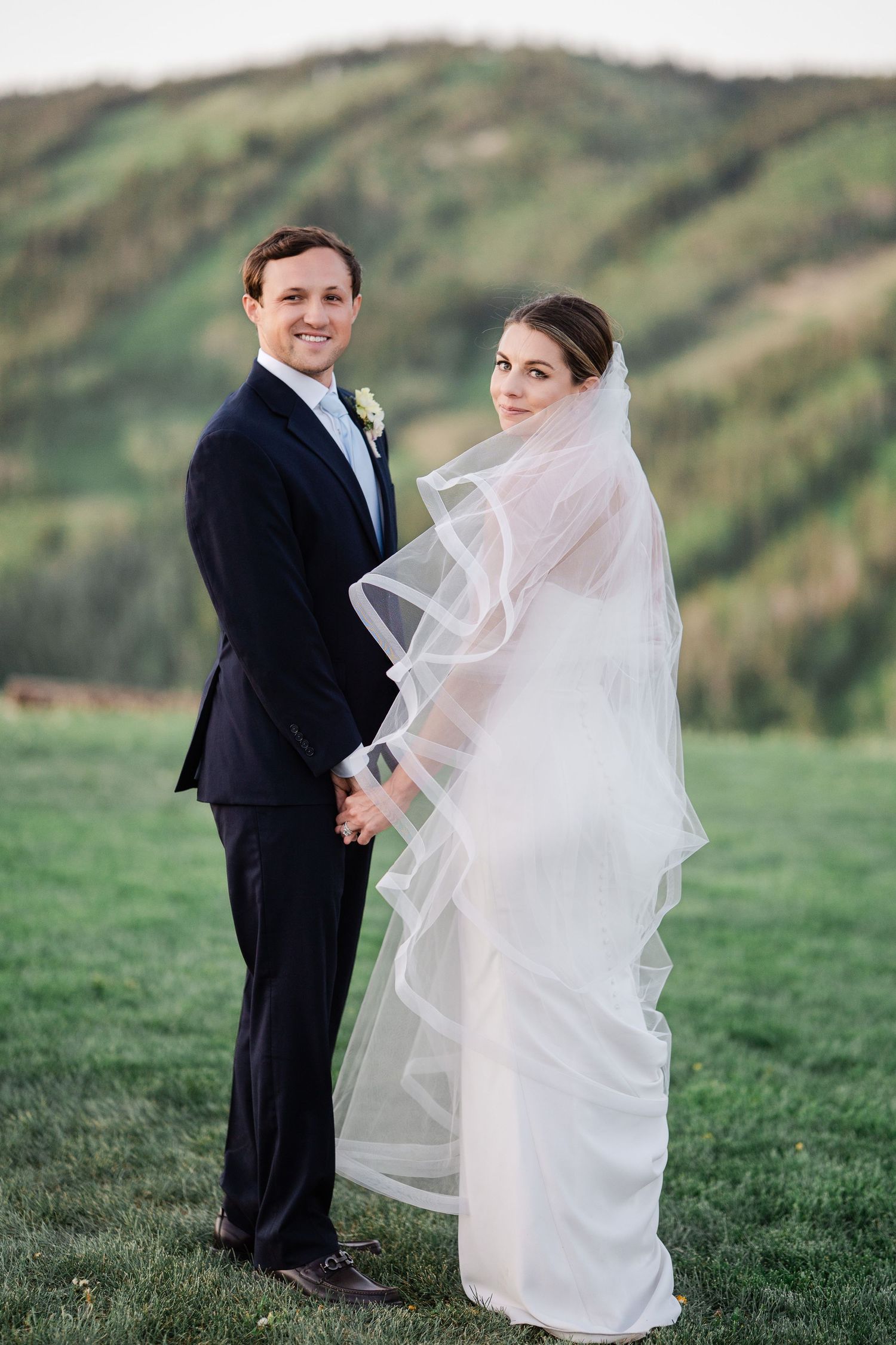 Bride and groom holding hands outdoors with rolling green hills backdrop, bride wearing flowing veil.
