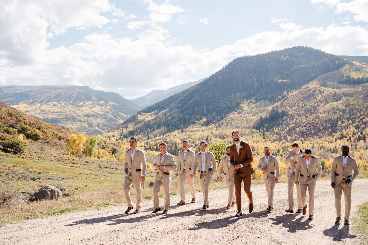 Groom and groomsmen in tan suits walk along a gravel road with colorful mountain valley backdrop.