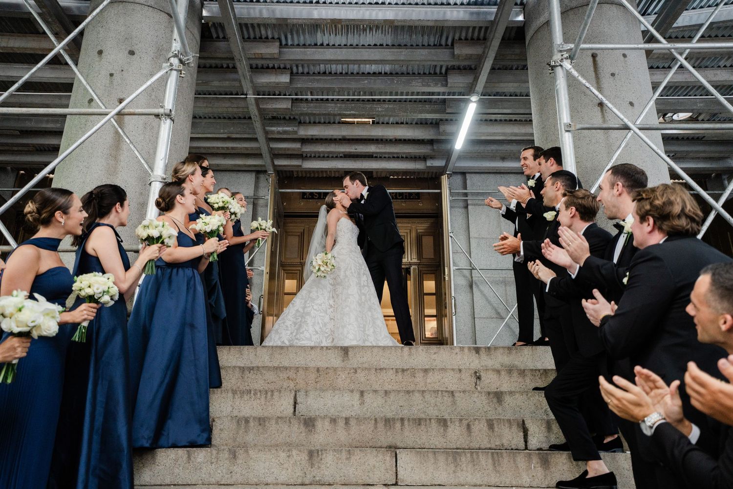 Wedding party celebrates couple kissing on grand staircase with bridesmaids in navy gowns.