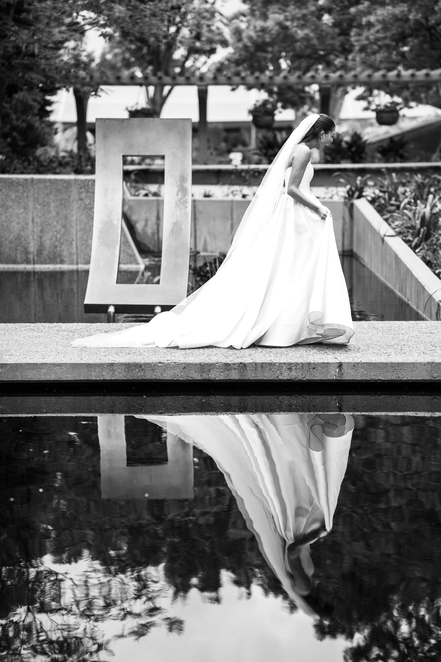 Black and white photo of bride in elegant gown reflected in still pool at outdoor venue.