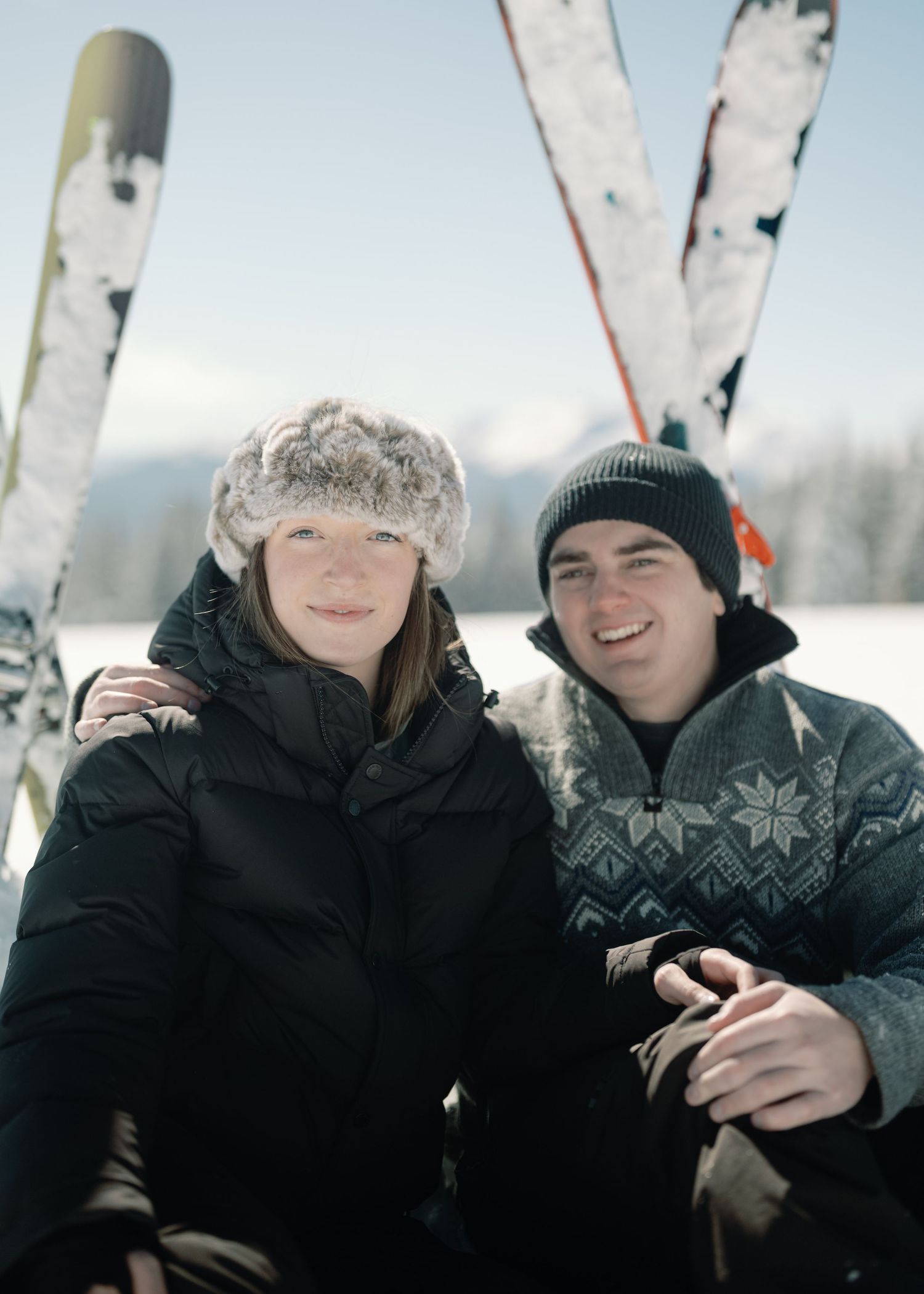 Smiling couple sitting together in snow with crossed skis and mountain backdrop on sunny day.