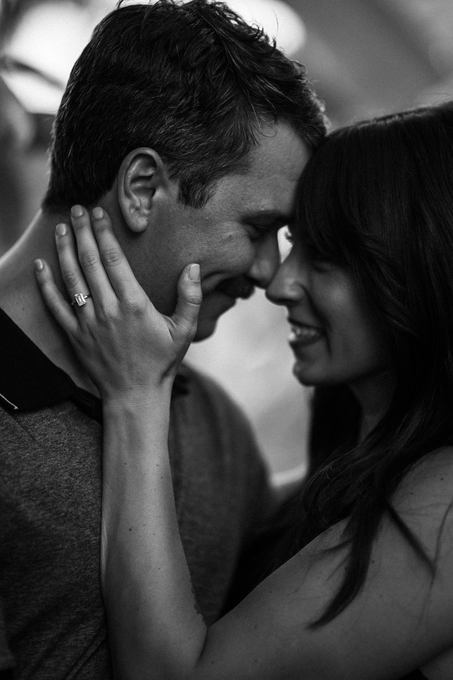 Romantic black and white close-up of engaged couple touching foreheads, woman showing diamond engagement ring.