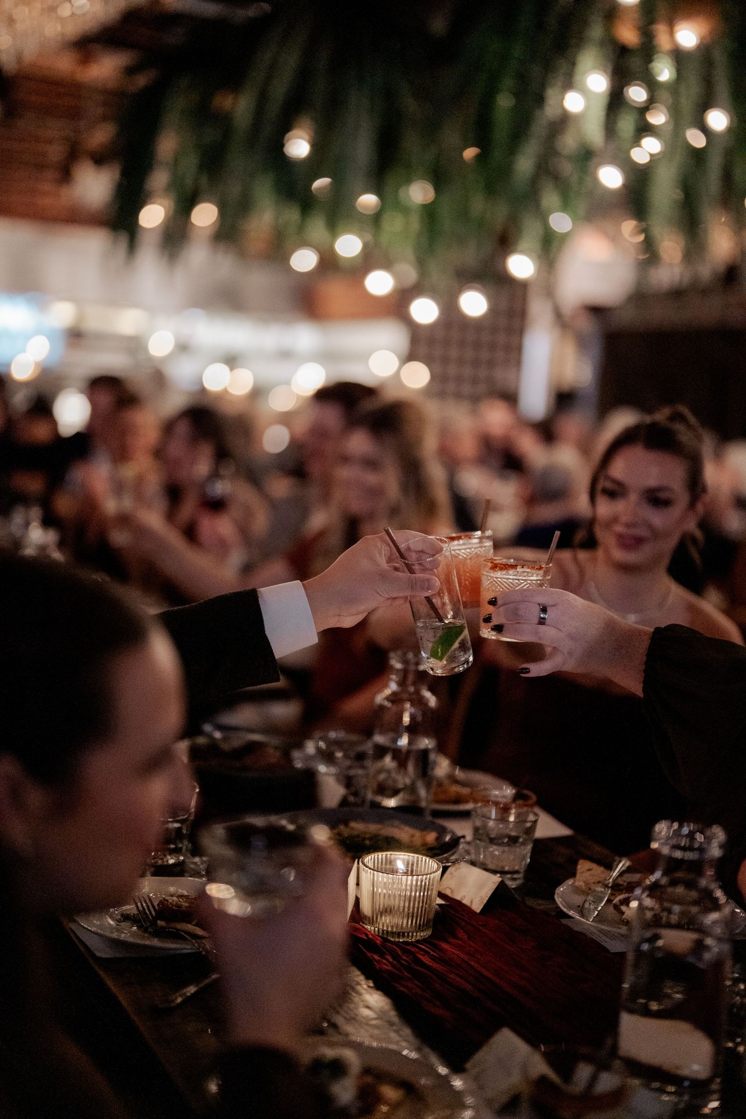 Wedding guests clink cocktail glasses together at candlelit reception table adorned with string lights and greenery.