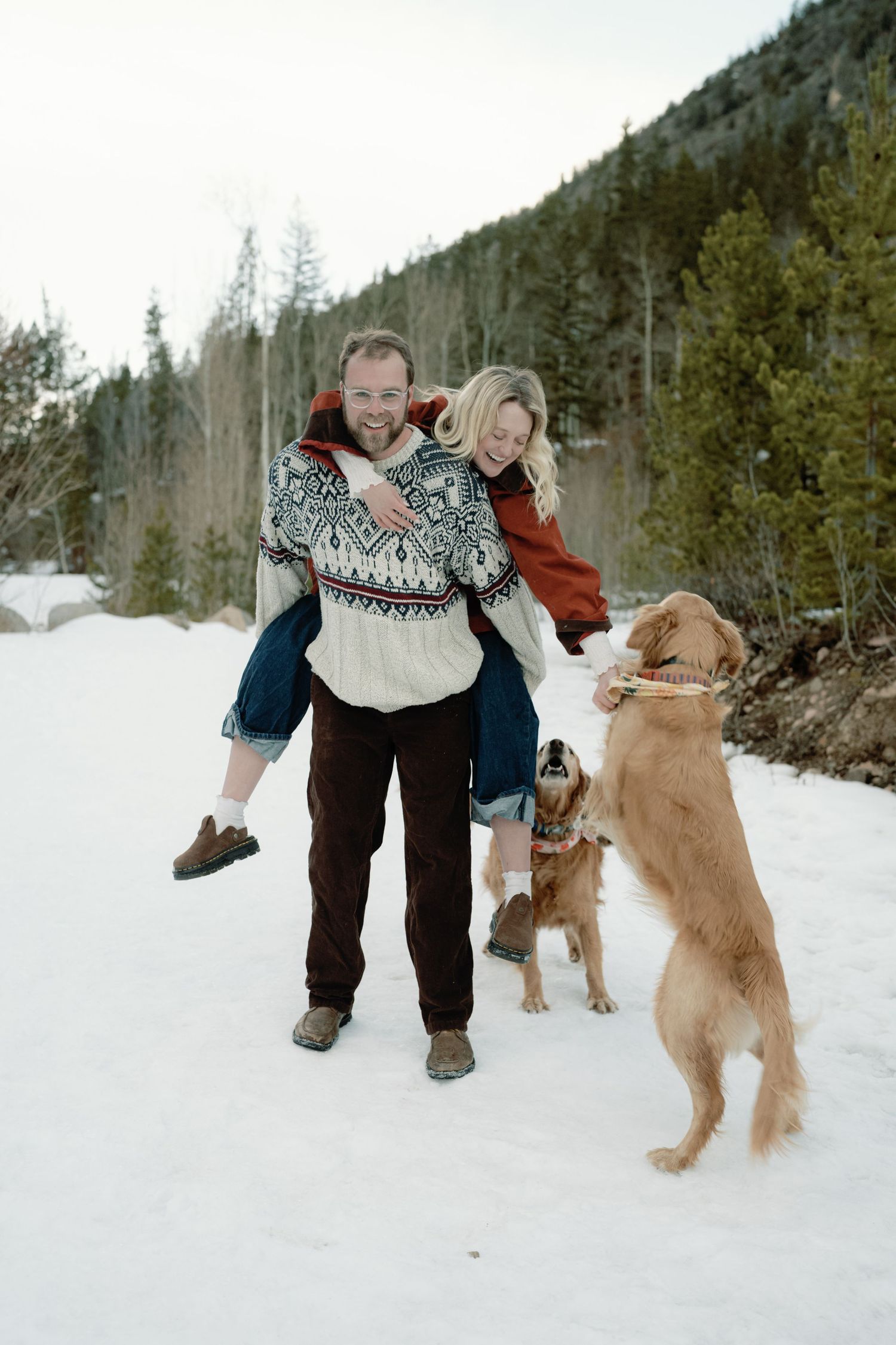 Couple laughing during piggyback ride in snow with two golden retrievers playing nearby in winter.