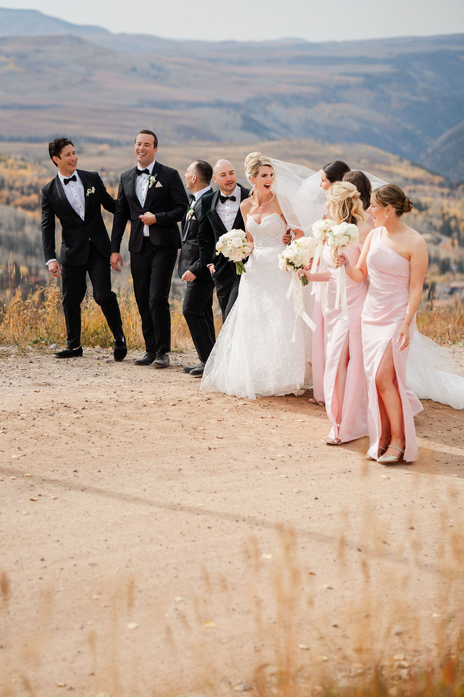 Wedding party walking together along mountain dirt path with golden autumn valley views in background.