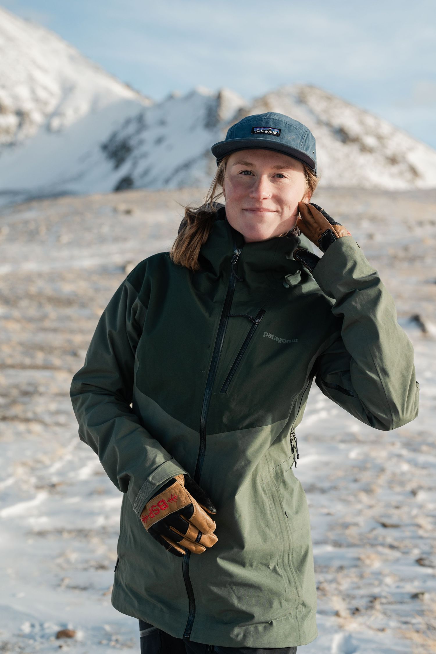 Woman in green jacket and cap holding gloves with snowy mountains in background on clear winter day.