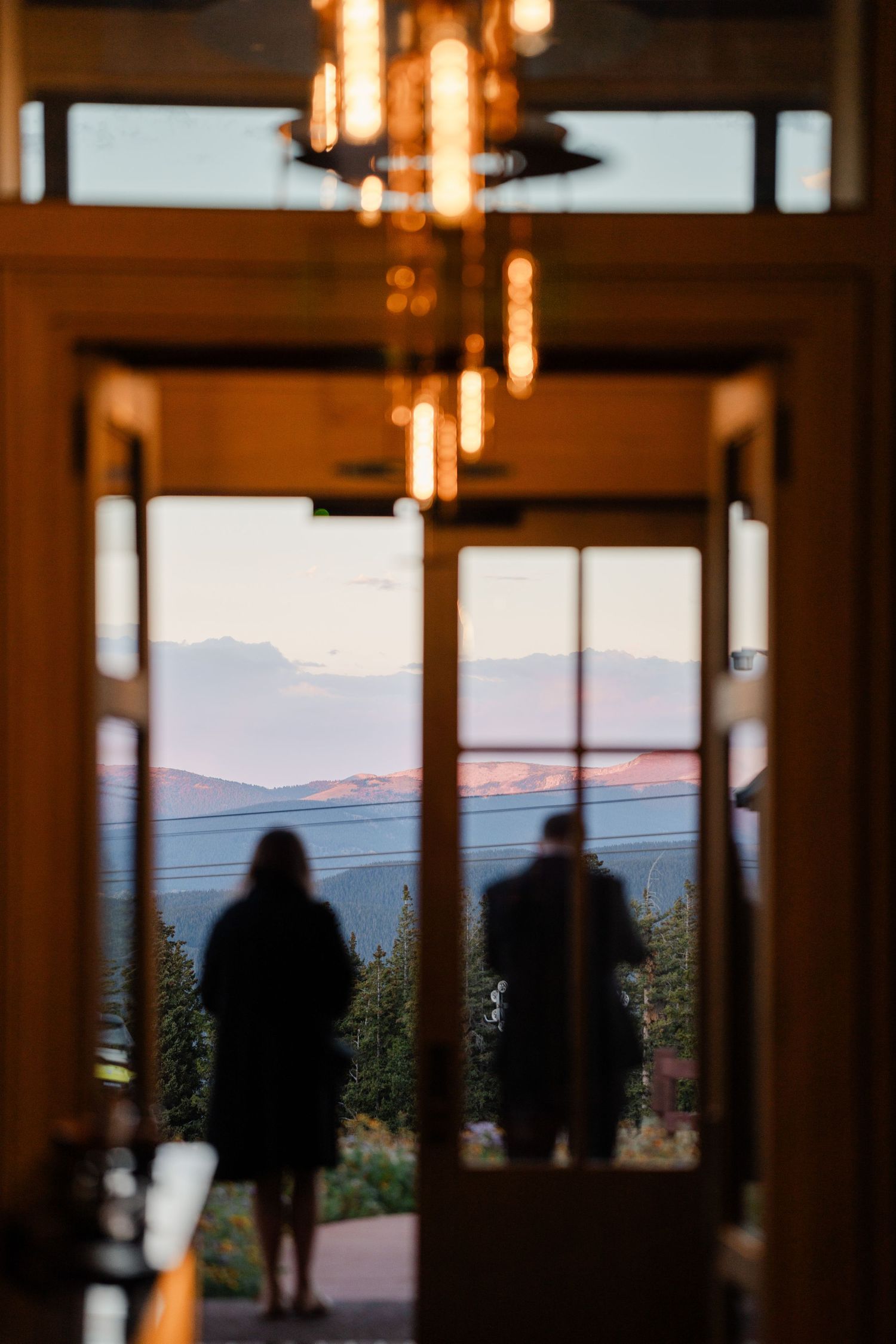 Silhouettes of couple standing at glass doors with mountain sunset view framed by warm chandelier light.