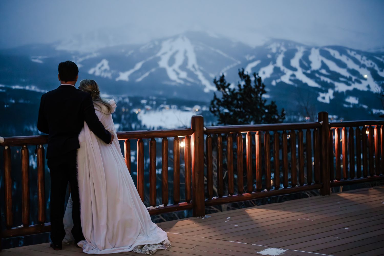 Bride and groom share a romantic moment on a wooden deck overlooking a snow-covered ski mountain at dusk.