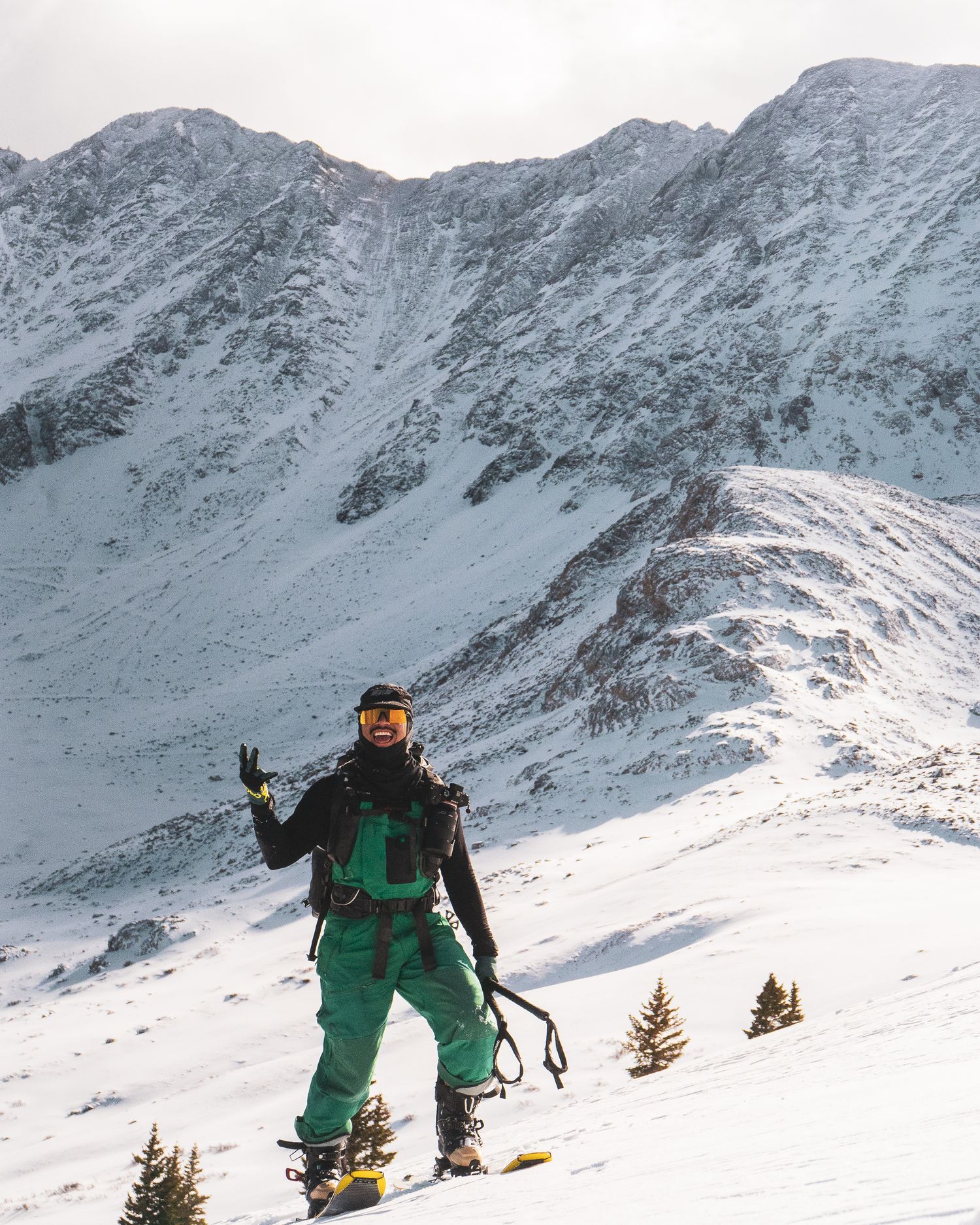 Snowboarder in green suit posing on snowy mountain slope with dramatic alpine peaks behind.