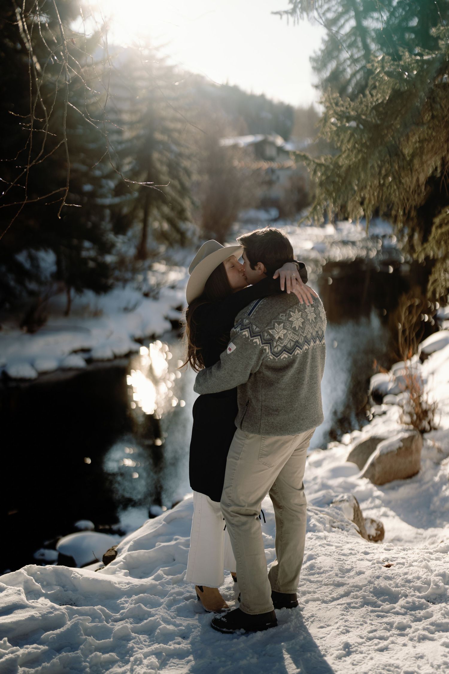 Couple embracing in a snowy winter forest with sunlight streaming through pine trees near a creek.