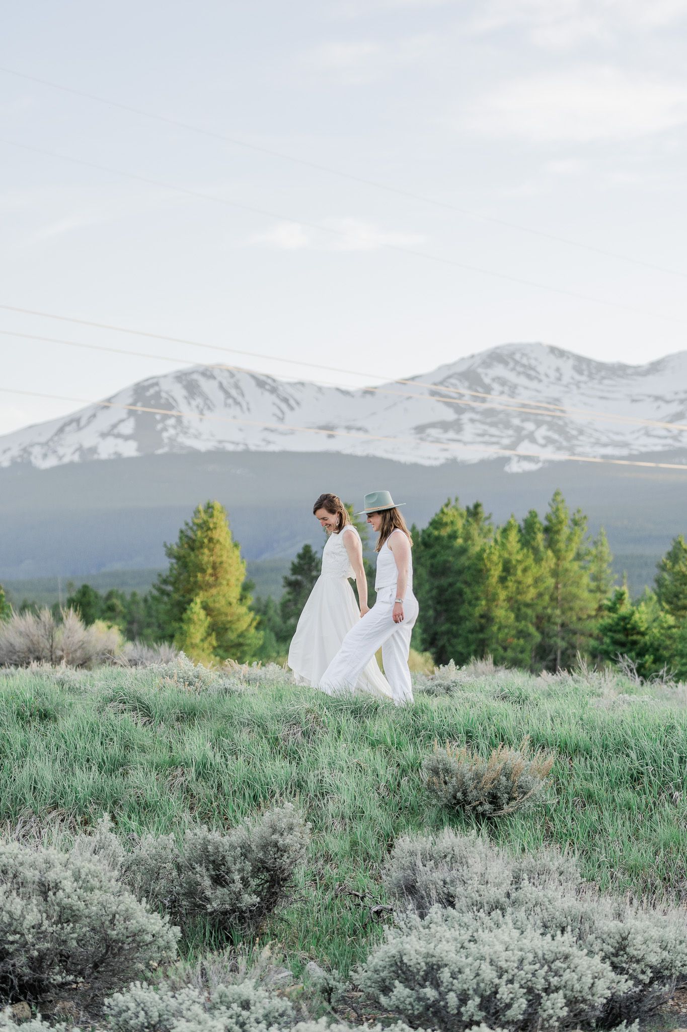 Two brides in white dresses walking through wildflower meadow with snow-capped mountains behind.