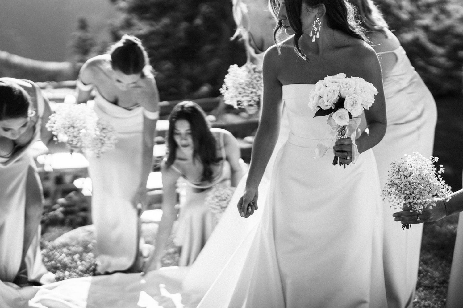 Bride in strapless white gown holding bouquet while bridesmaids adjust her dress outdoors in black and white.