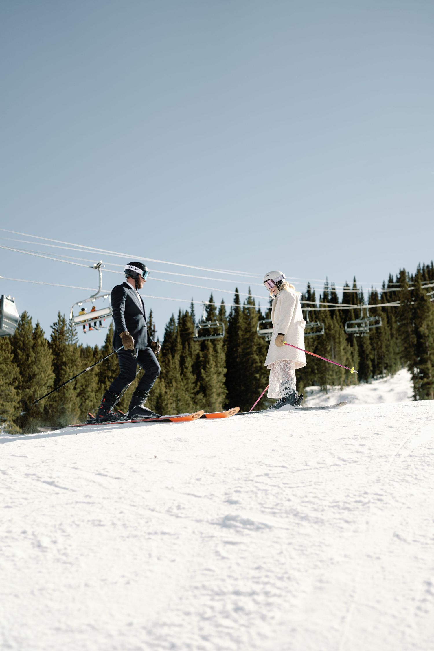 Bride and groom skiing together on a sunny mountain slope with ski lifts overhead.