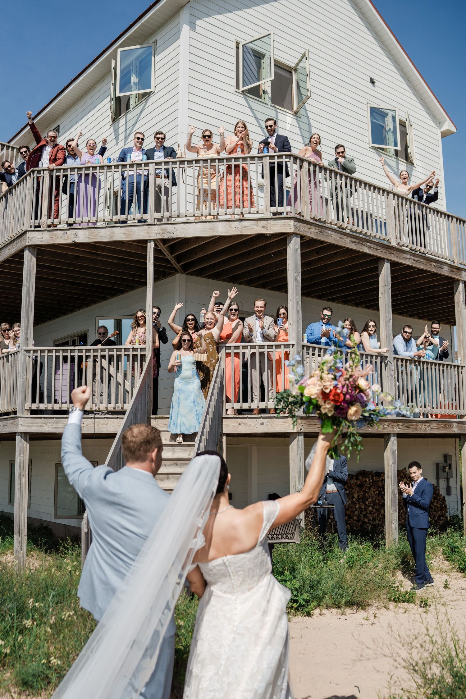 Newlyweds celebrate as wedding guests cheer from multi-level deck of white beachside house on sunny day.