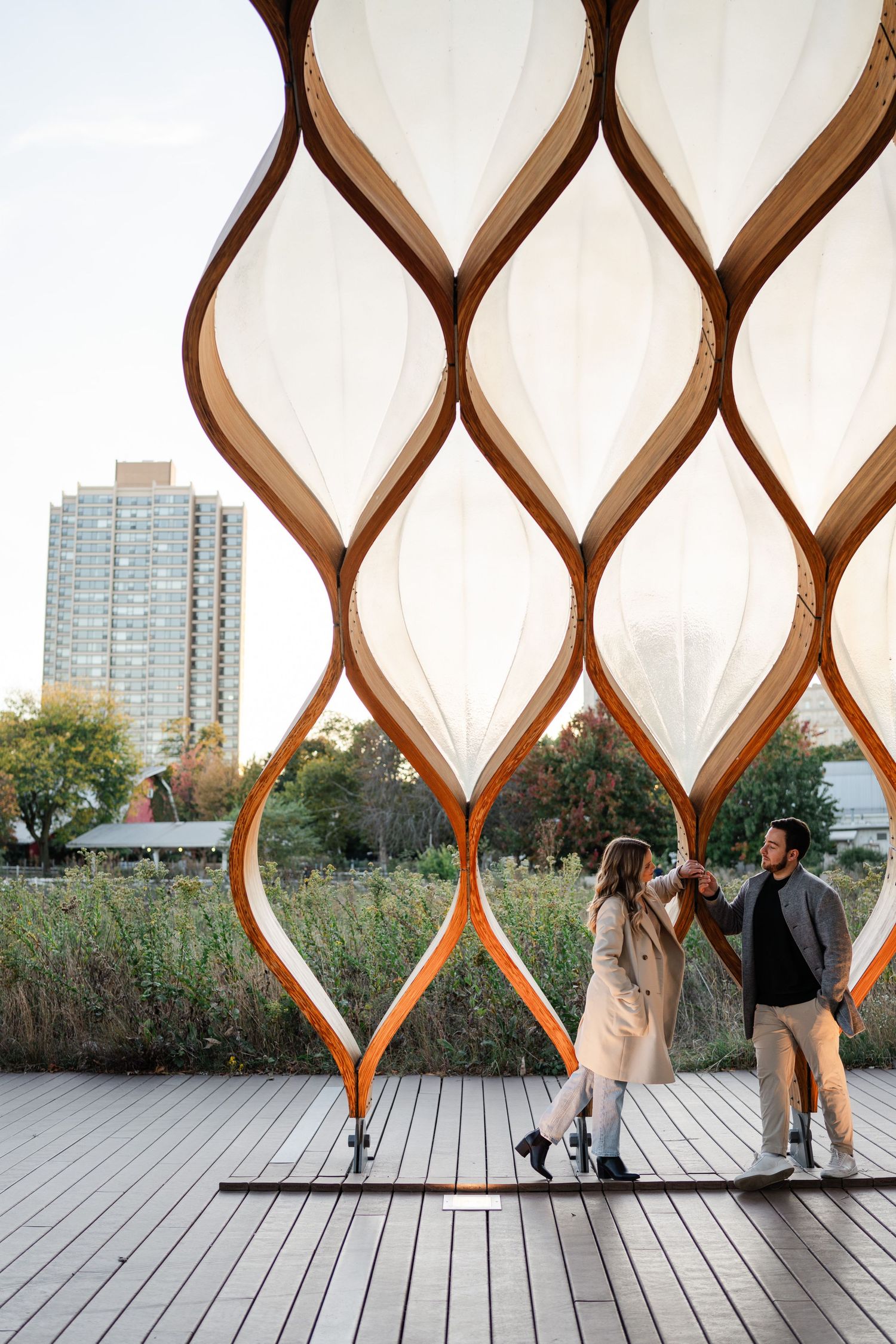 Couple exploring a stunning wooden lattice pavilion installation with translucent panels against a city skyline backdrop.