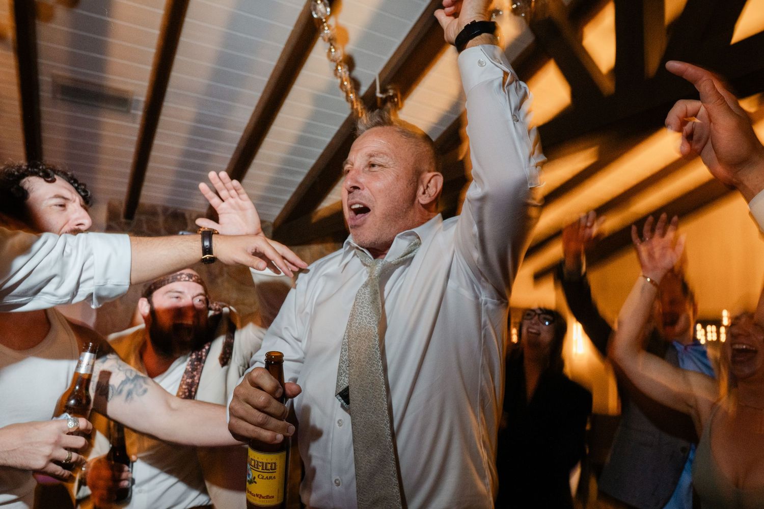 Excited wedding guest celebrating at reception, holding beer bottle while cheering with friends in rustic venue.