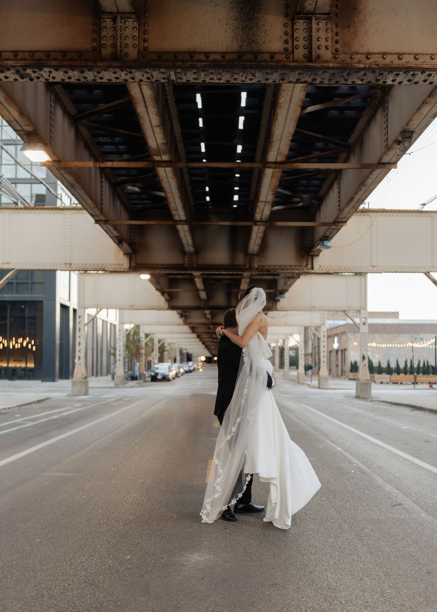 Bride and groom sharing romantic embrace on empty urban street under elevated train tracks.
